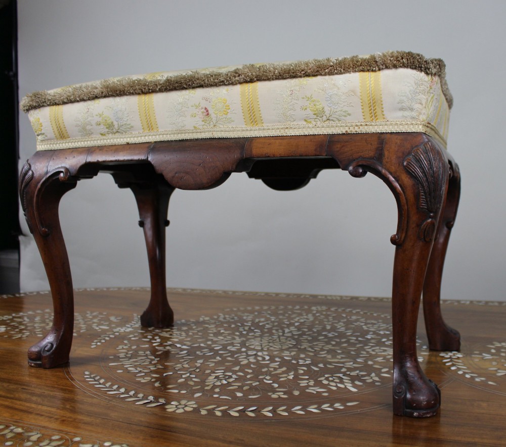 A low walnut footstool featuring carved cabriole legs and a cream and yellow floral striped fabric upholstered seat with tassel trim. View IV