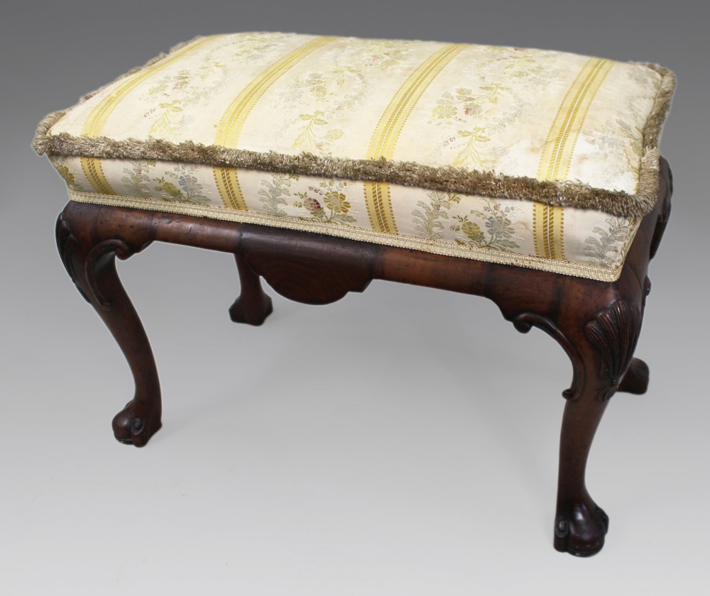 A low walnut footstool featuring carved cabriole legs and a cream and yellow floral striped fabric upholstered seat with tassel trim. View VI