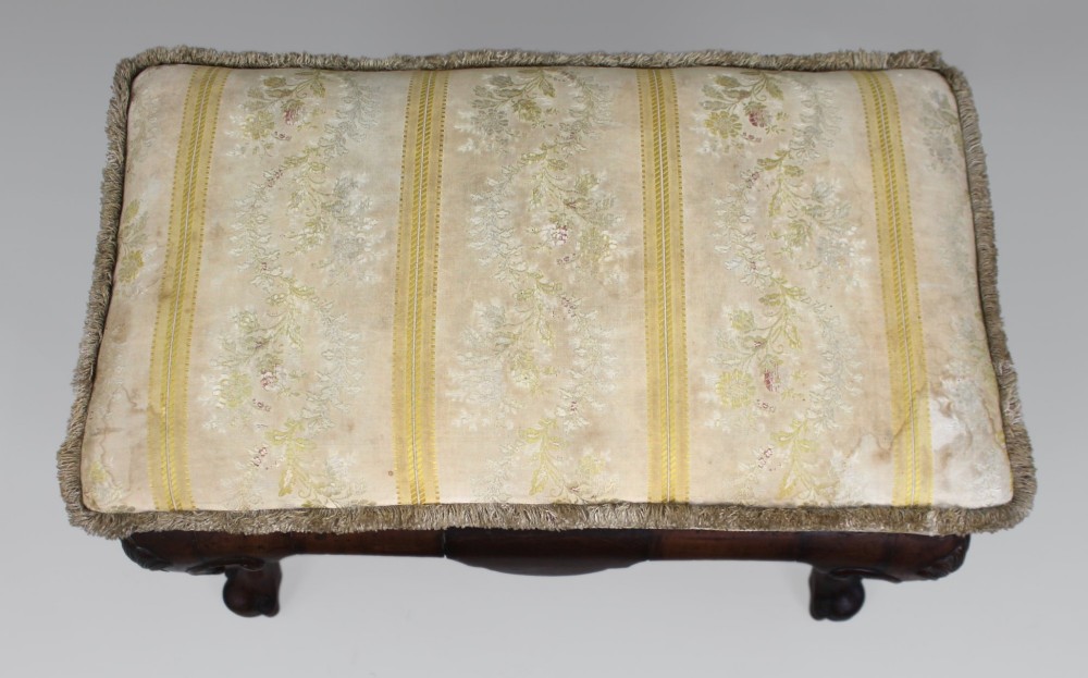 A low walnut footstool featuring carved cabriole legs and a cream and yellow floral striped fabric upholstered seat with tassel trim. View X