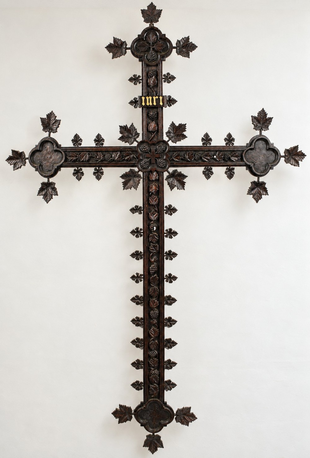 A nineteenth-century dark wood Black Forest wall cross detailed with carved vine leaves, grape clusters, trefoil terminals, and gilded INRI lettering.