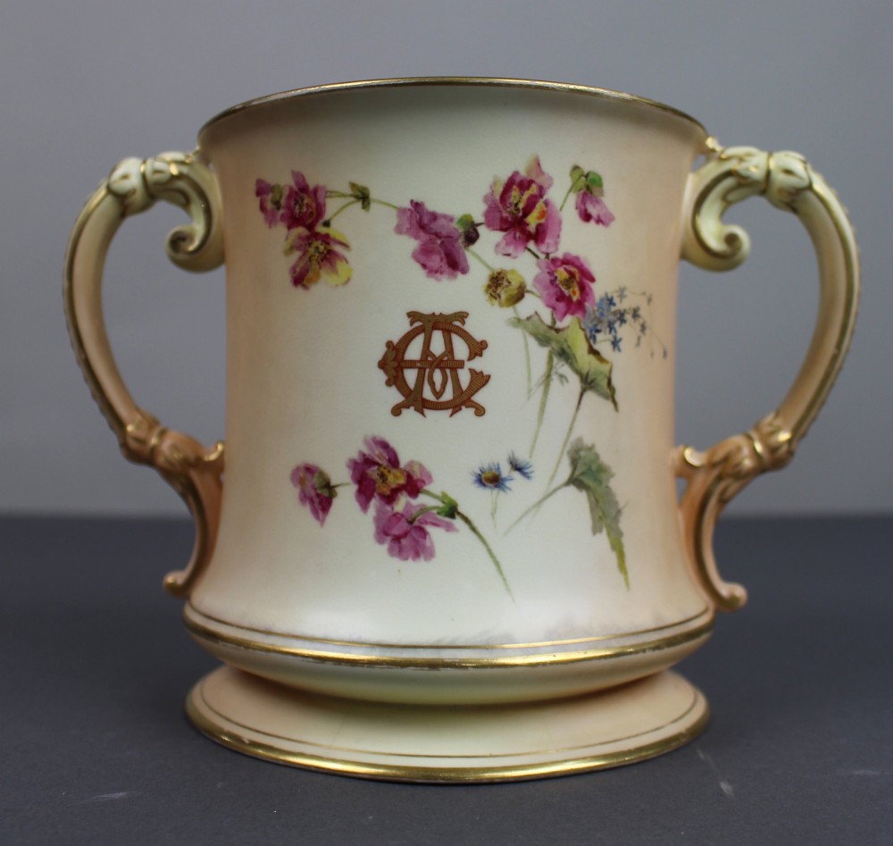 An Edwardian Royal Worcester porcelain loving cup featuring twin gilded scroll handles, a cream blush ground, hand-painted floral motifs, and a central gilt monogram.