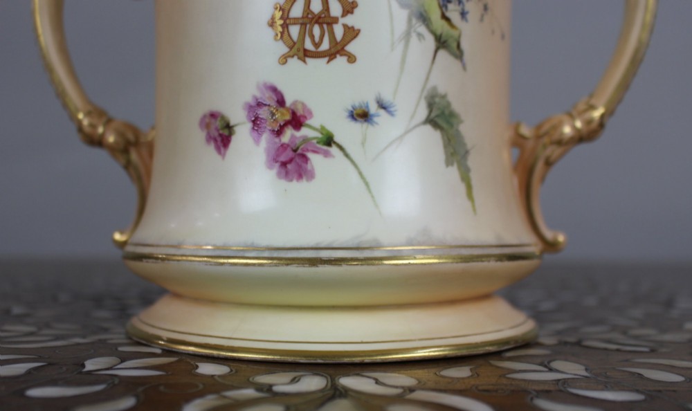 An Edwardian Royal Worcester porcelain loving cup featuring twin gilded scroll handles, a cream blush ground, hand-painted floral motifs, and a central gilt monogram. View V