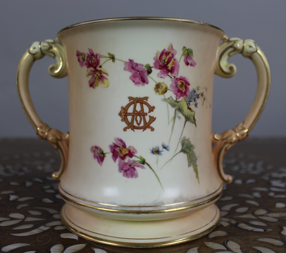 An Edwardian Royal Worcester porcelain loving cup featuring twin gilded scroll handles, a cream blush ground, hand-painted floral motifs, and a central gilt monogram. View VI