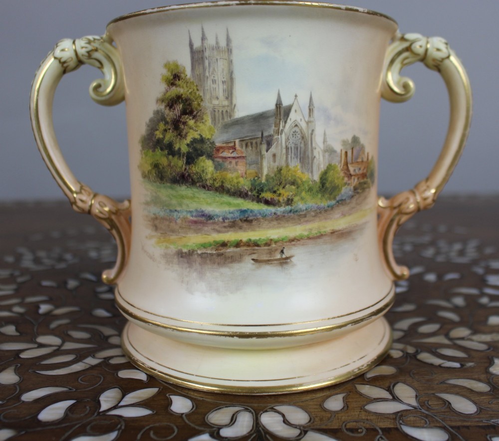 An Edwardian Royal Worcester porcelain loving cup featuring twin gilded scroll handles, a cream blush ground, hand-painted floral motifs, and a central gilt monogram. View VII