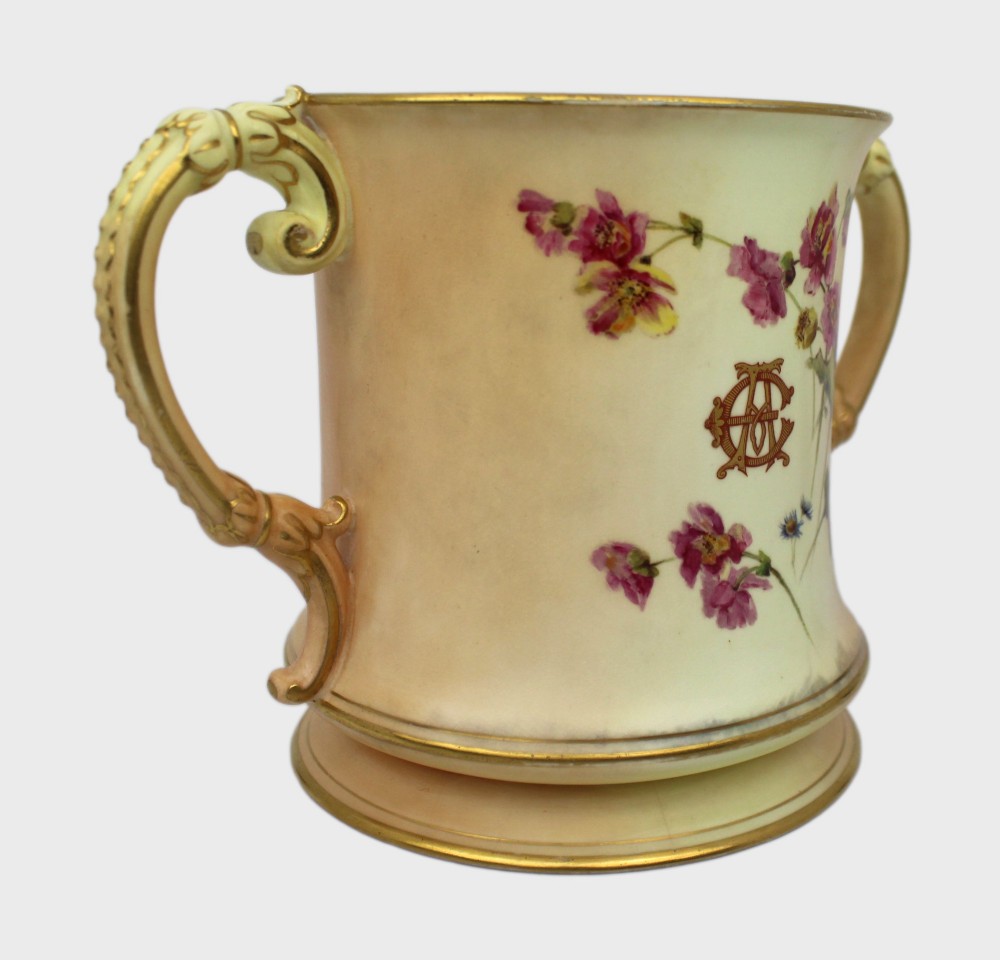 An Edwardian Royal Worcester porcelain loving cup featuring twin gilded scroll handles, a cream blush ground, hand-painted floral motifs, and a central gilt monogram. View XII