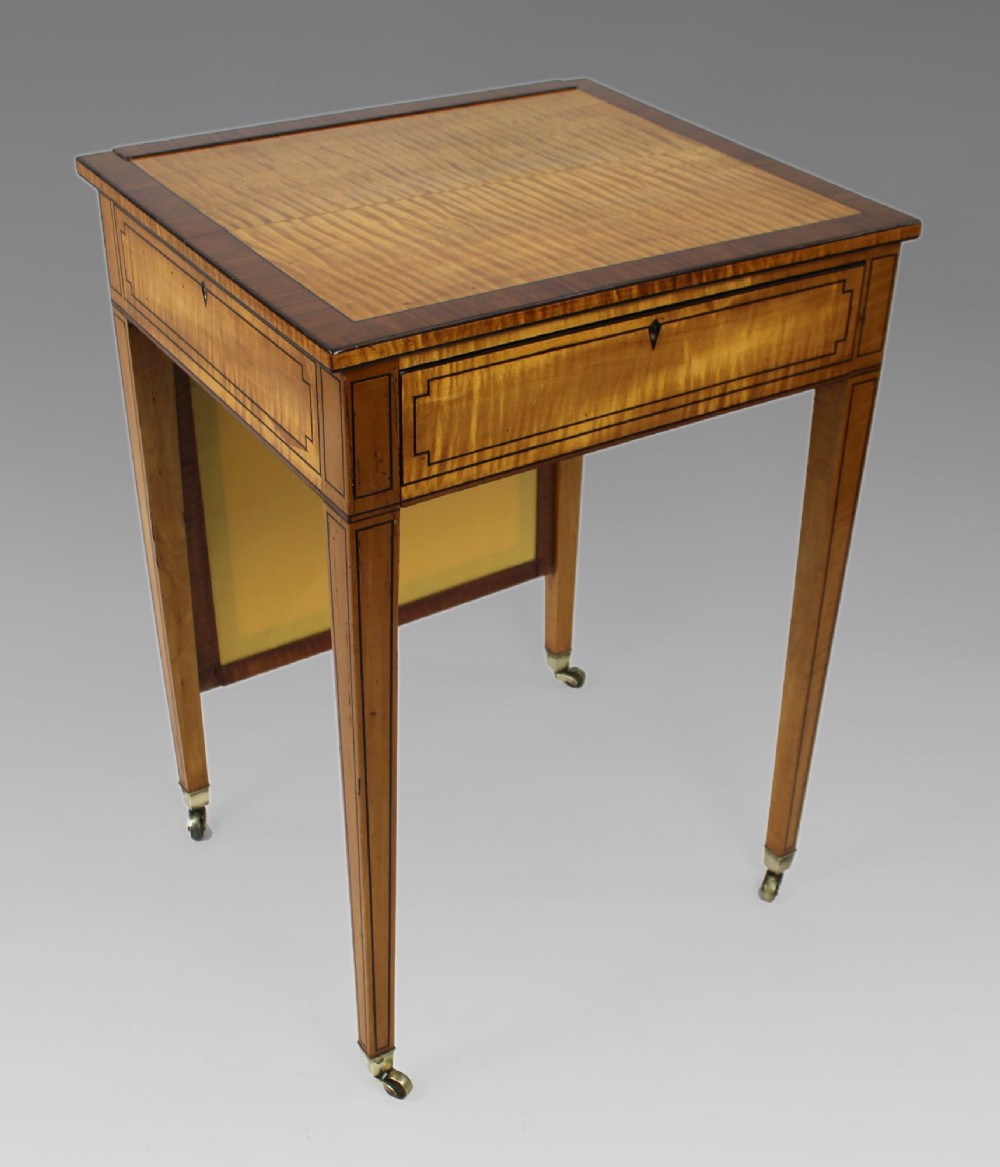 A George III Sheraton satinwood writing table featuring dark wood crossbanding, line inlay, a single drawer, square tapered legs, and brass castors.