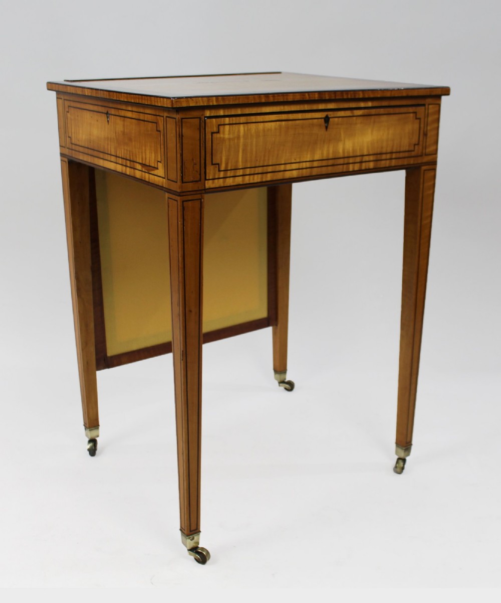 A George III Sheraton satinwood writing table featuring dark wood crossbanding, line inlay, a single drawer, square tapered legs, and brass castors. View II