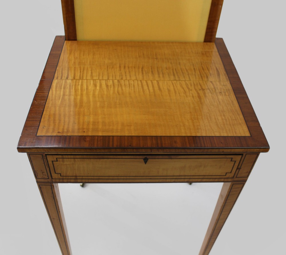 A George III Sheraton satinwood writing table featuring dark wood crossbanding, line inlay, a single drawer, square tapered legs, and brass castors. View IV