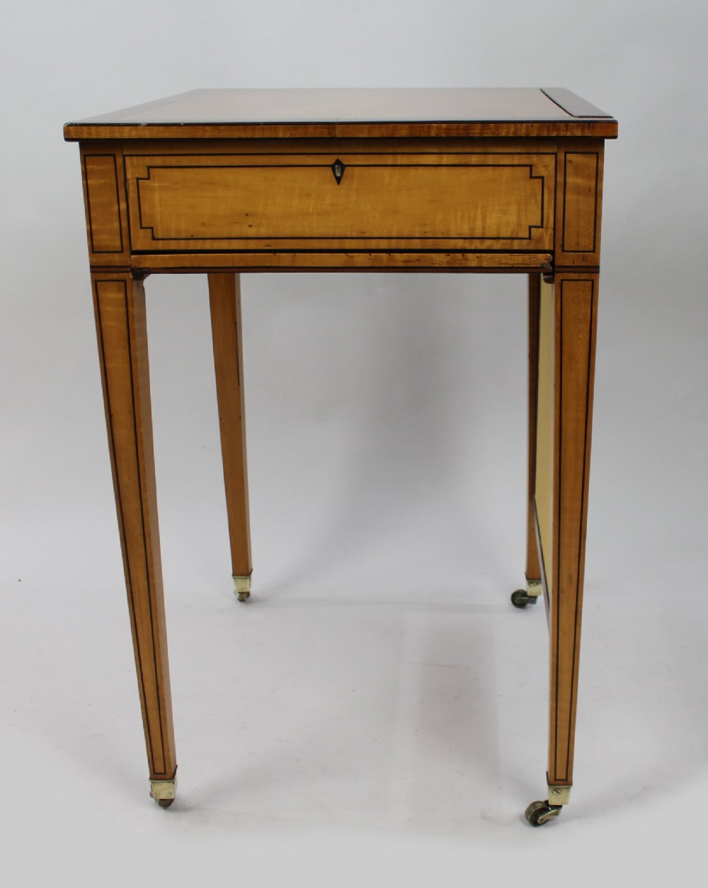 A George III Sheraton satinwood writing table featuring dark wood crossbanding, line inlay, a single drawer, square tapered legs, and brass castors. View XI
