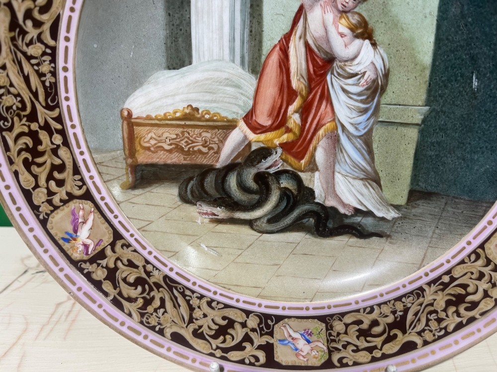 A circular 19th-century Meissen porcelain cabinet plate featuring a central hand-painted classical scene framed by a dark burgundy rim with intricate gilded scrollwork and cherub cartouches. View V