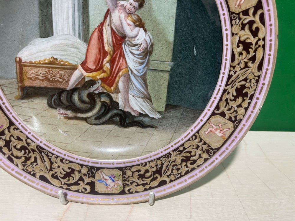 A circular 19th-century Meissen porcelain cabinet plate featuring a central hand-painted classical scene framed by a dark burgundy rim with intricate gilded scrollwork and cherub cartouches. View VI