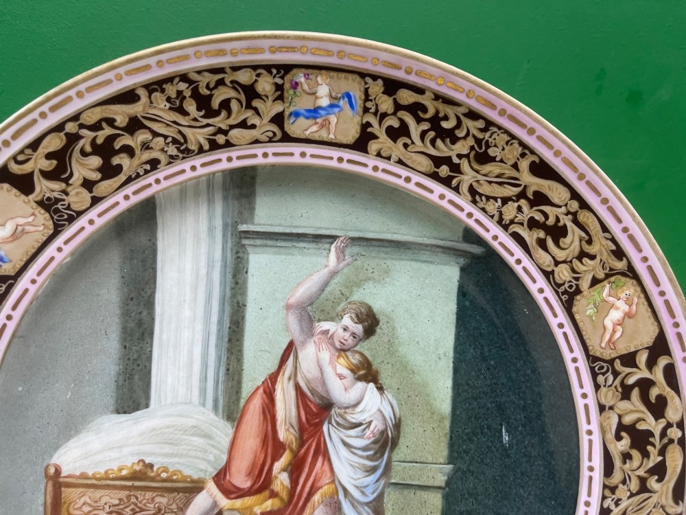 A circular 19th-century Meissen porcelain cabinet plate featuring a central hand-painted classical scene framed by a dark burgundy rim with intricate gilded scrollwork and cherub cartouches. View VII