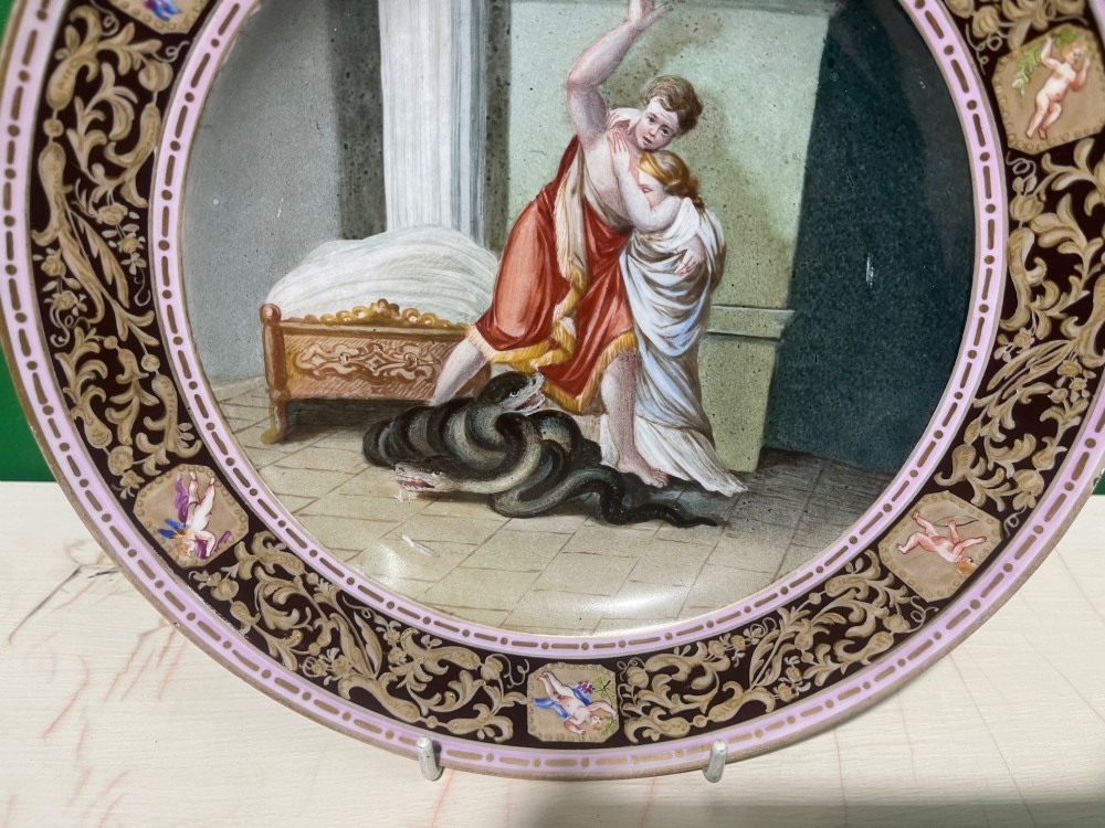 A circular 19th-century Meissen porcelain cabinet plate featuring a central hand-painted classical scene framed by a dark burgundy rim with intricate gilded scrollwork and cherub cartouches. View X