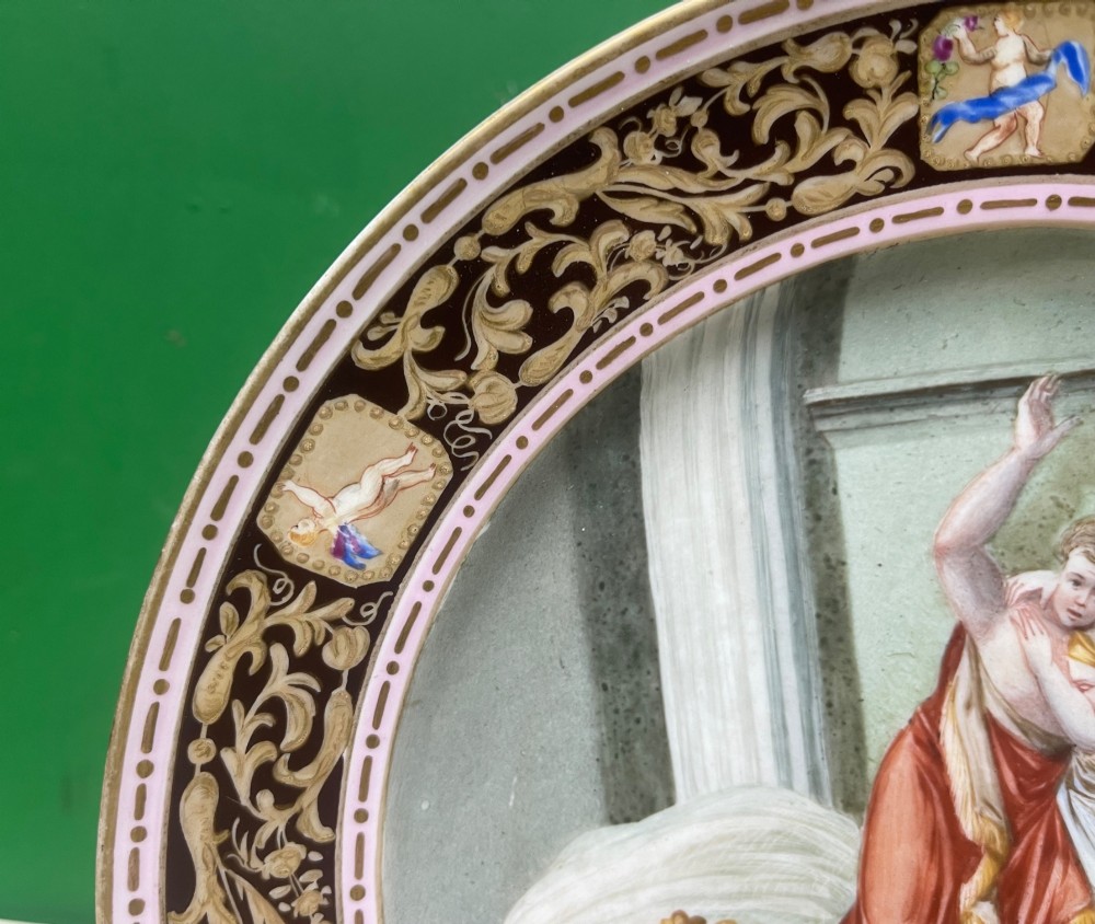 A circular 19th-century Meissen porcelain cabinet plate featuring a central hand-painted classical scene framed by a dark burgundy rim with intricate gilded scrollwork and cherub cartouches. View XII