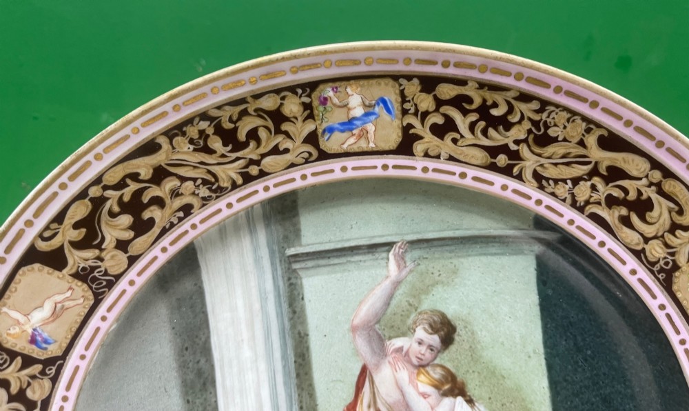 A circular 19th-century Meissen porcelain cabinet plate featuring a central hand-painted classical scene framed by a dark burgundy rim with intricate gilded scrollwork and cherub cartouches. View XIII