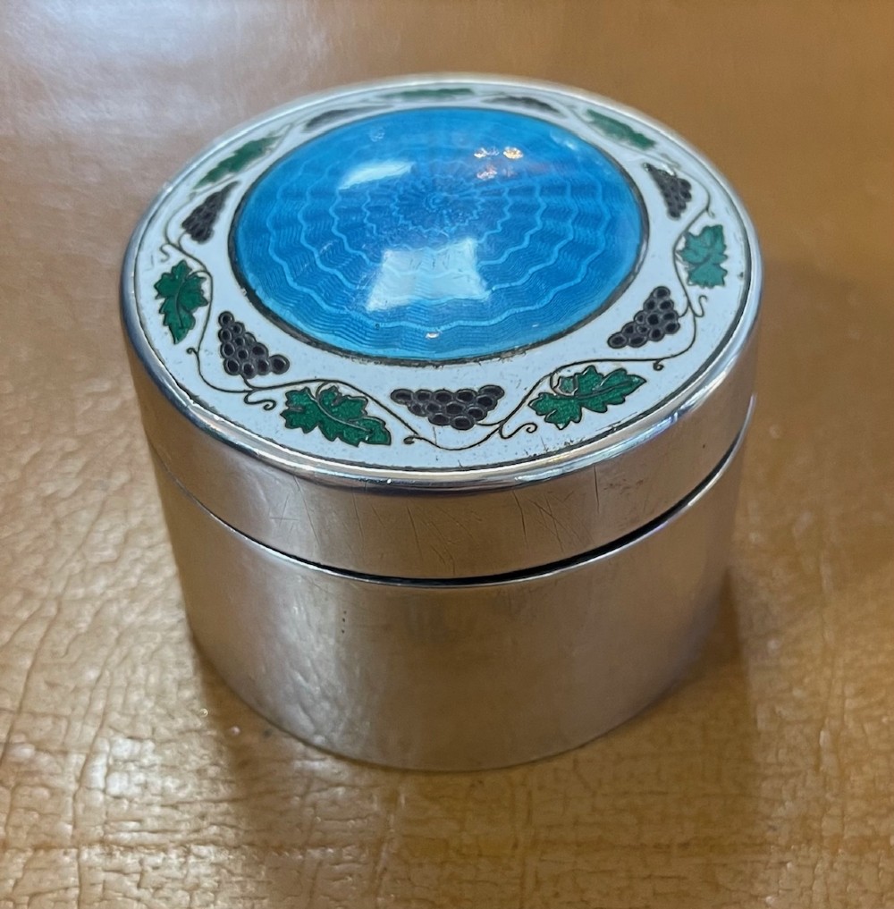 A circular Edwardian silver box lid featuring a blue engine-turned guilloche enamel centre surrounded by a white enamel border with green foliage and dark grape motifs. View II