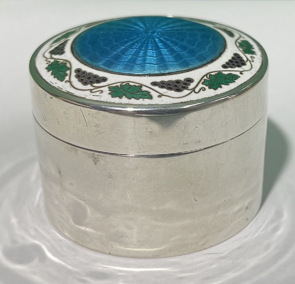 A circular Edwardian silver box lid featuring a blue engine-turned guilloche enamel centre surrounded by a white enamel border with green foliage and dark grape motifs. View VI