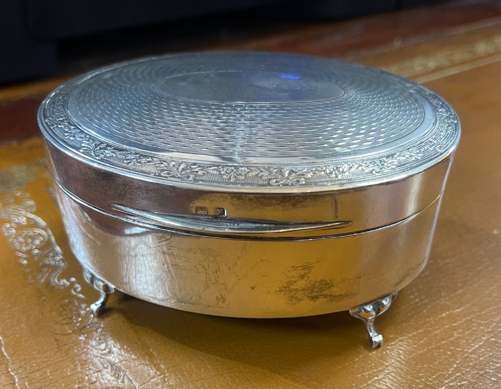 An oval sterling silver footed trinket casket featuring an engine-turned top, floral engraved lid border, and four scroll feet.