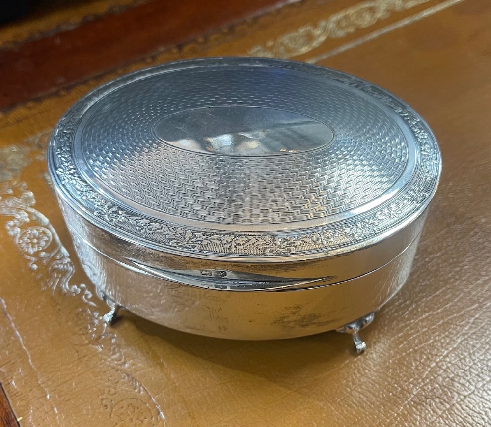 An oval sterling silver footed trinket casket featuring an engine-turned top, floral engraved lid border, and four scroll feet. View II