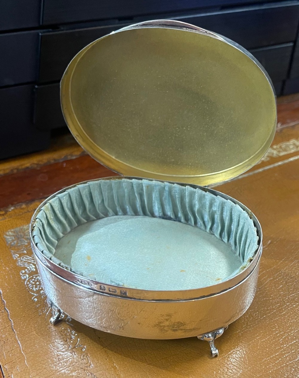 An oval sterling silver footed trinket casket featuring an engine-turned top, floral engraved lid border, and four scroll feet. View III