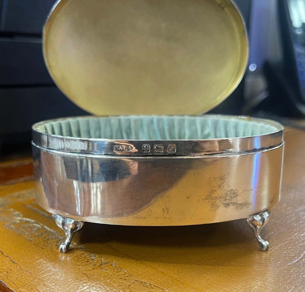 An oval sterling silver footed trinket casket featuring an engine-turned top, floral engraved lid border, and four scroll feet. View IV