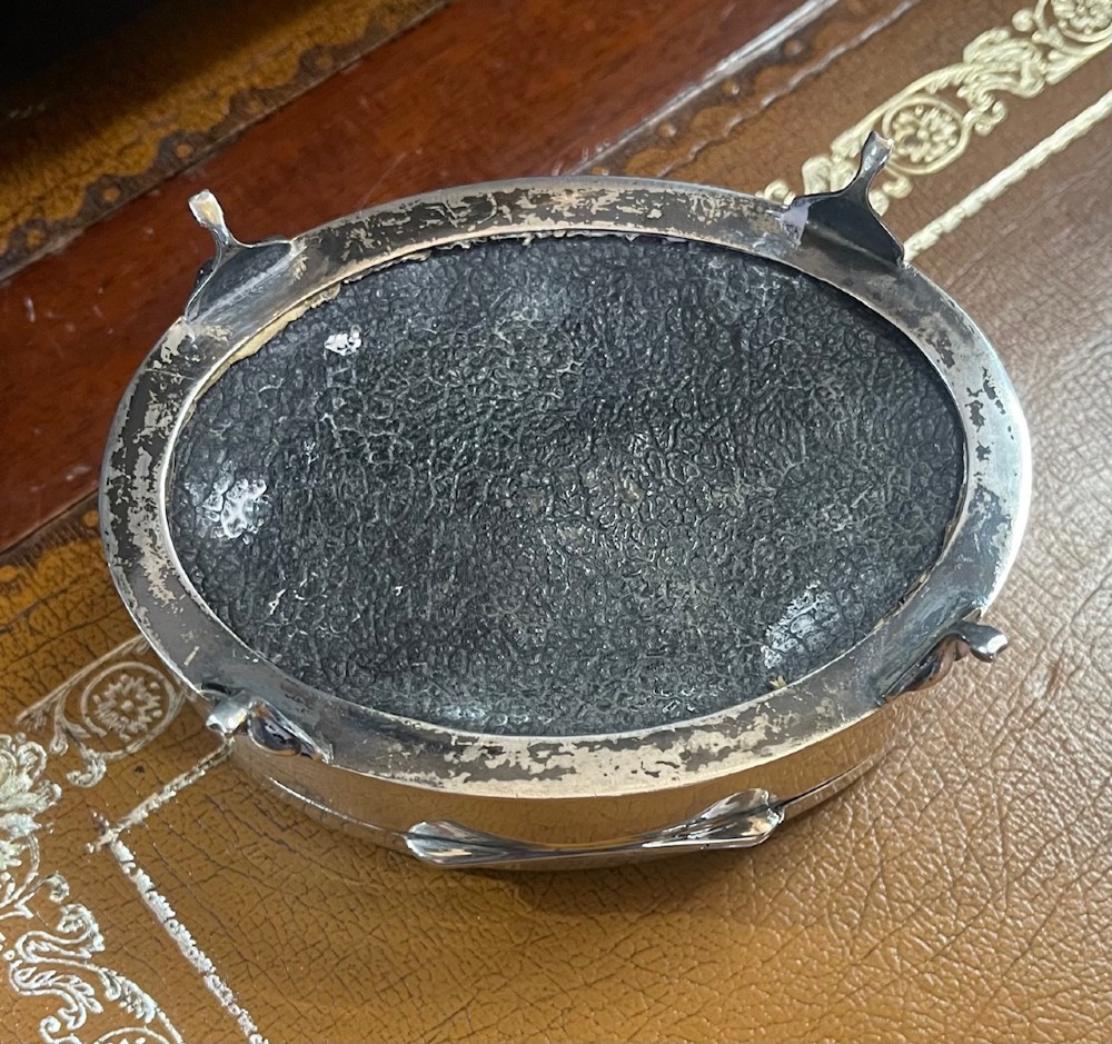 An oval sterling silver footed trinket casket featuring an engine-turned top, floral engraved lid border, and four scroll feet. View V