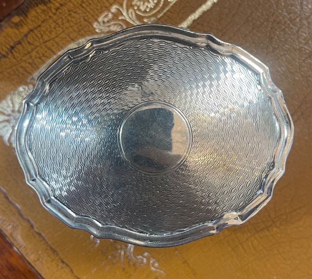 An oval sterling silver trinket box featuring an engine-turned wave pattern on the lid and resting on four small feet. View V