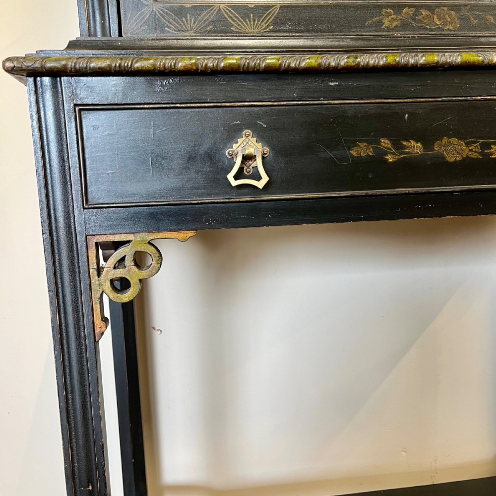 An ebonised Aesthetic Movement display cabinet featuring a glazed upper section with geometric glazing bars, hand-painted gilt motifs, a drawer, and slim legs with a bottom tier shelf. View VI