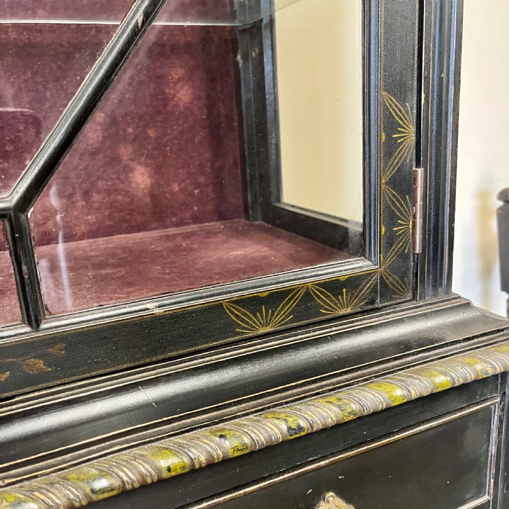 An ebonised Aesthetic Movement display cabinet featuring a glazed upper section with geometric glazing bars, hand-painted gilt motifs, a drawer, and slim legs with a bottom tier shelf. View X