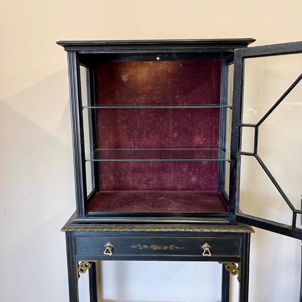 An ebonised Aesthetic Movement display cabinet featuring a glazed upper section with geometric glazing bars, hand-painted gilt motifs, a drawer, and slim legs with a bottom tier shelf. View XII