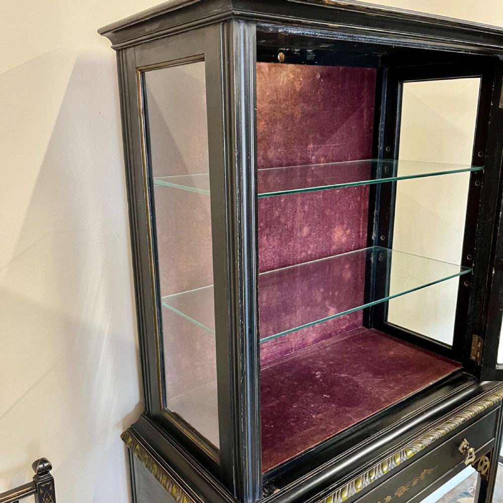 An ebonised Aesthetic Movement display cabinet featuring a glazed upper section with geometric glazing bars, hand-painted gilt motifs, a drawer, and slim legs with a bottom tier shelf. View XIV