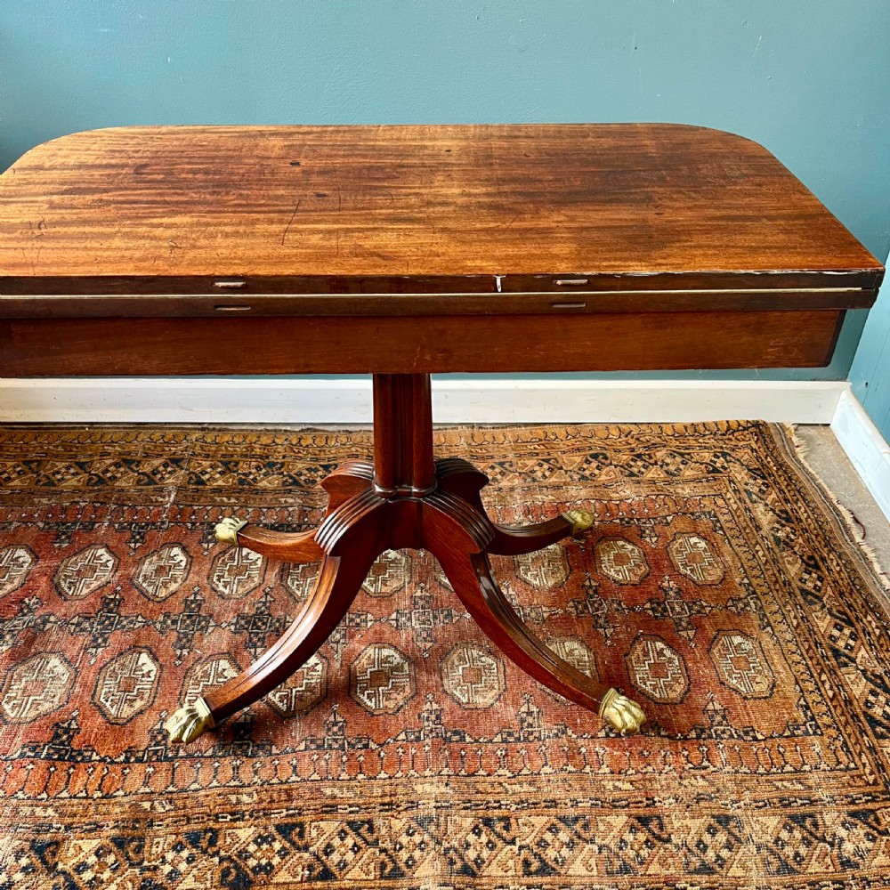 A 19th-century mahogany fold-over card table with a carved frieze, central pedestal, and splayed quadripartite legs ending in brass lion paw castors. View VII