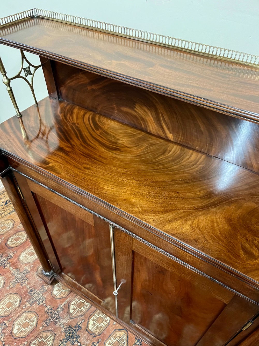 A 19th-century mahogany chiffonier featuring an upper shelf with a brass gallery, twin panelled cupboard doors flanked by turned side columns, resting on bun feet. View VI