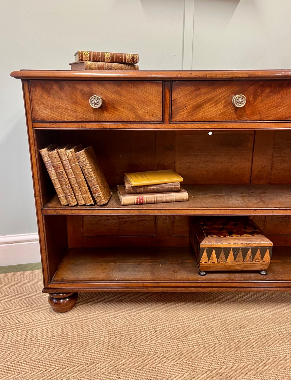 A low 19th-century mahogany bookcase featuring three small drawers above open shelving and supported by turned feet. View II