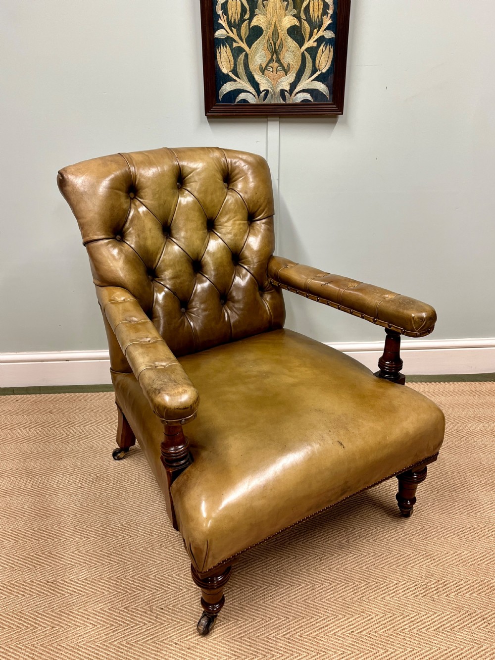 A 19th-century deep-buttoned tan leather armchair featuring padded armrests, brass studding, turned dark mahogany front legs, and castors.