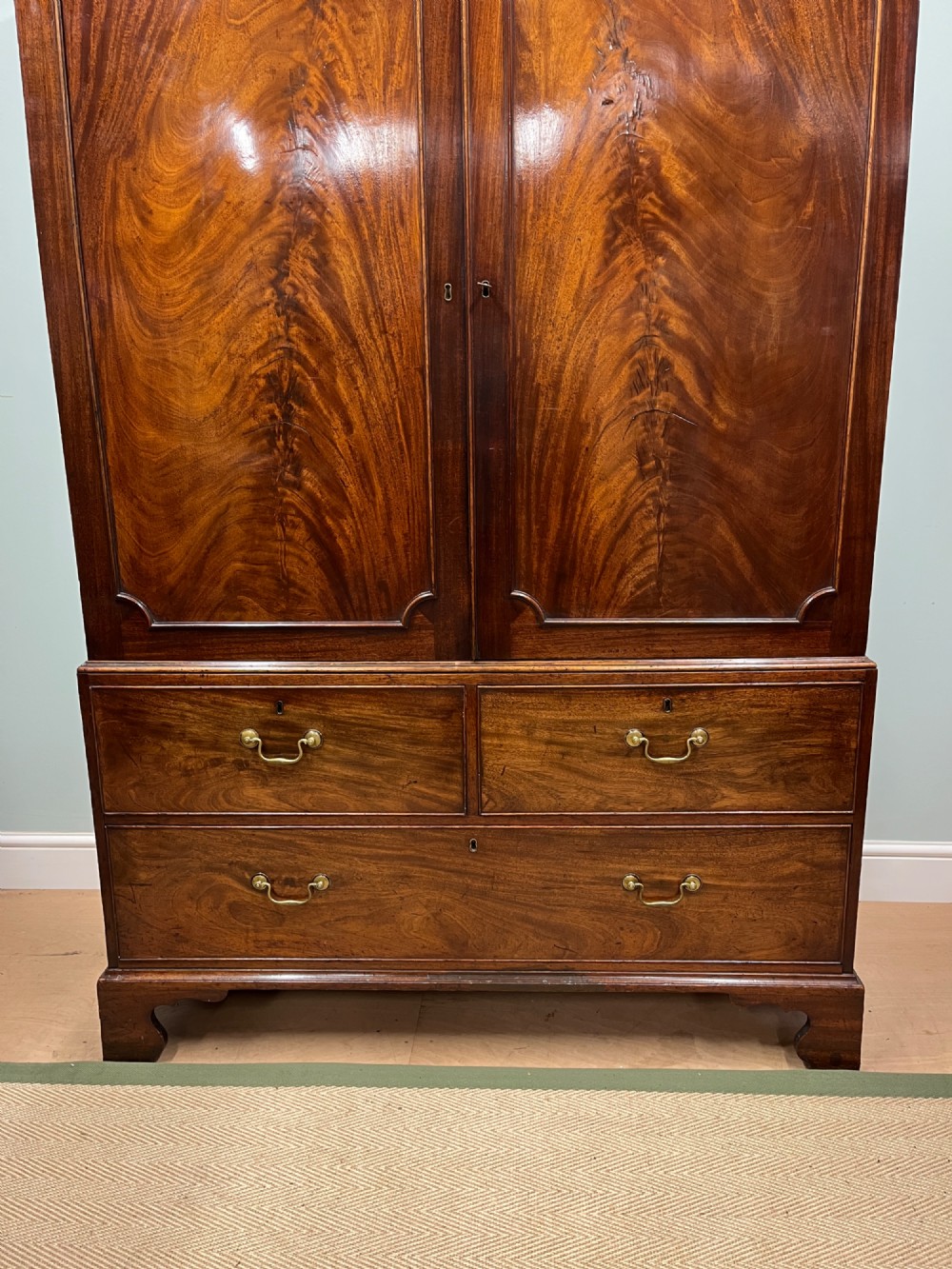 A nineteenth-century flame mahogany linen press featuring panelled cupboard doors above three lower drawers with brass swan-neck handles. View II