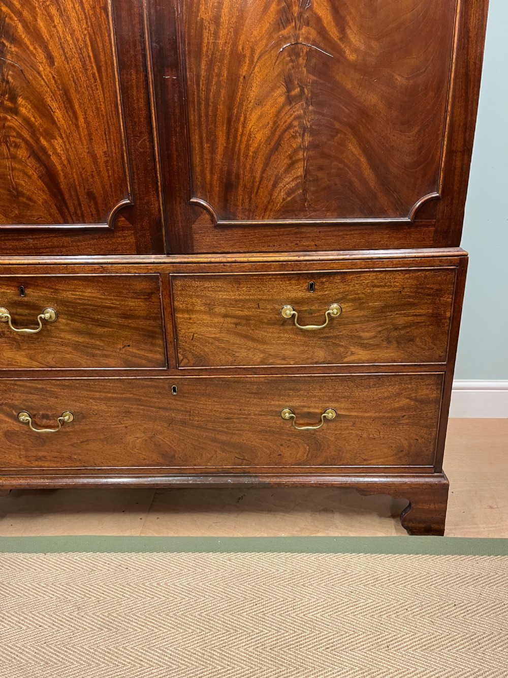 A nineteenth-century flame mahogany linen press featuring panelled cupboard doors above three lower drawers with brass swan-neck handles. View IV