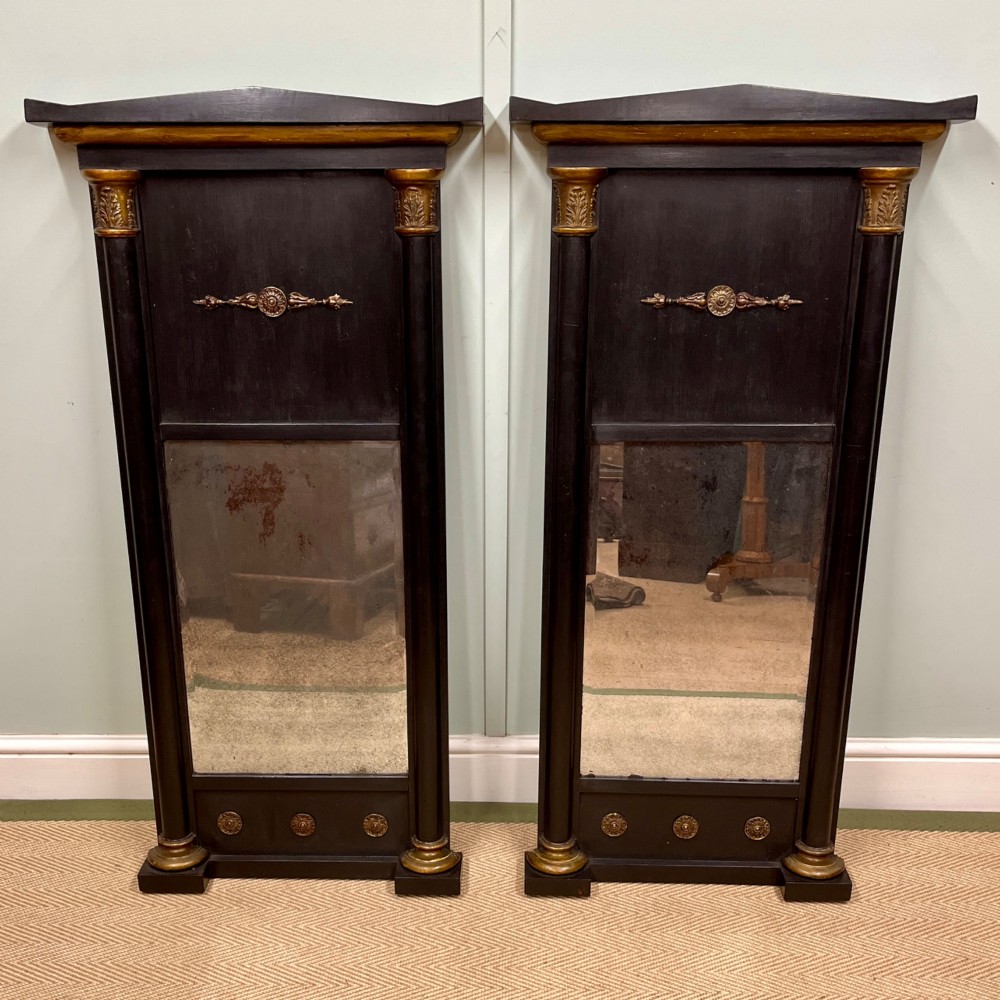 A pair of tall Empire style ebonised pier mirrors featuring gilt column capitals, architectural tops, and classical metal appliques.