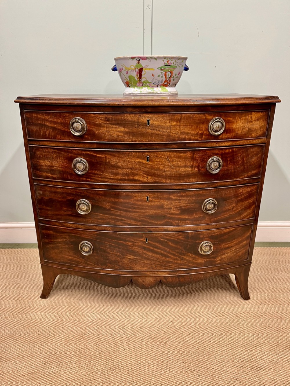 An antique Regency bow-fronted mahogany chest of drawers with four graduated drawers, brass ring handles, and splayed bracket feet.