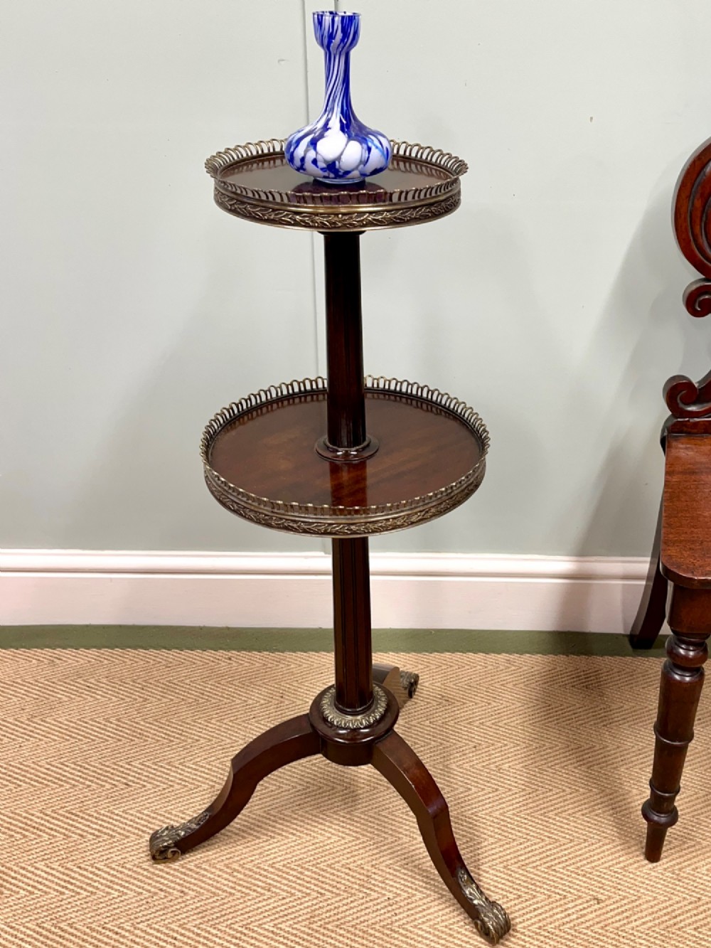 A two-tier dark mahogany dumb waiter featuring pierced brass galleries, a fluted central stem, and a tripod base with brass feet.