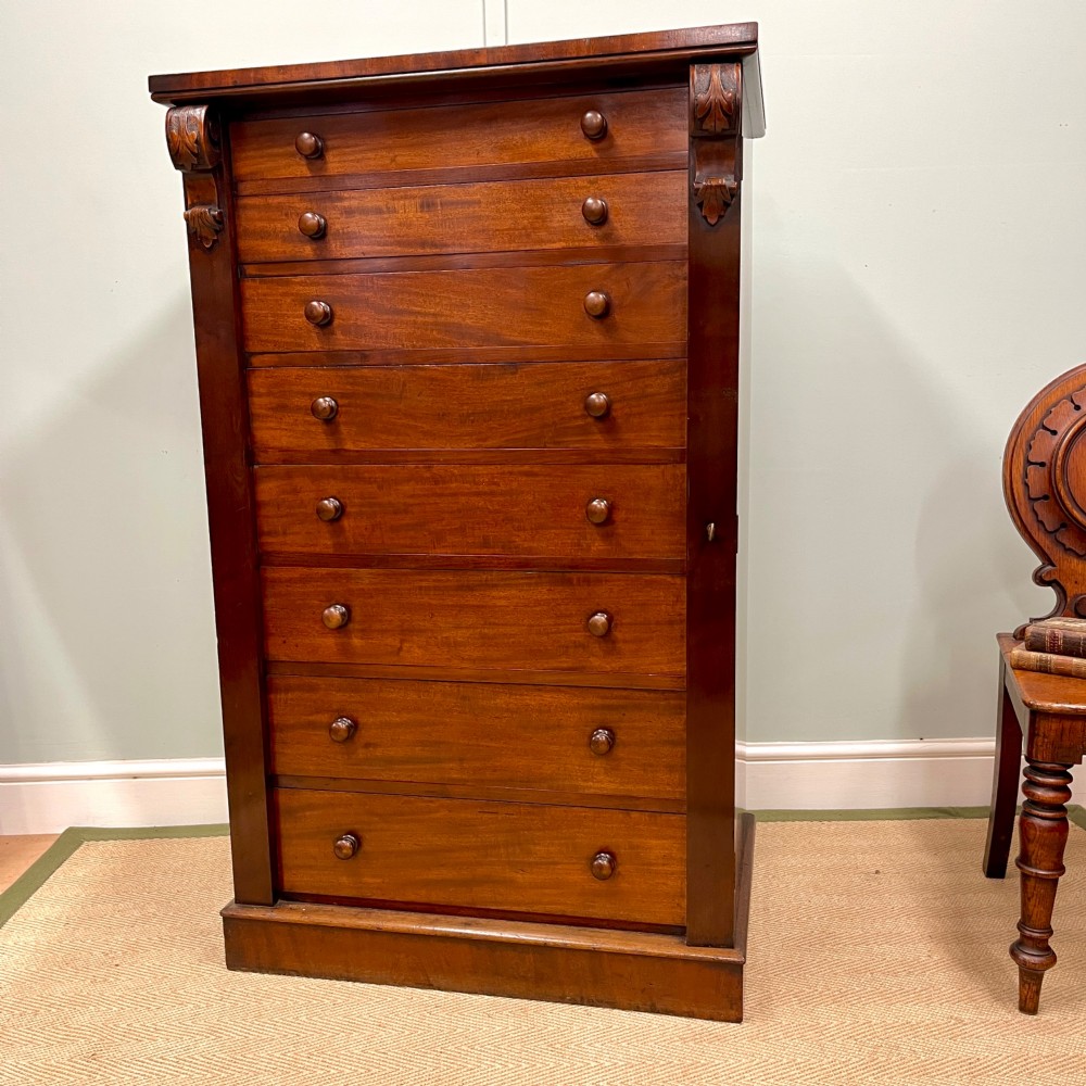 A tall Victorian mahogany Wellington chest featuring eight graduated drawers with turned wooden knobs, carved corbels at the top, and a side hinged locking flap on a plinth base.