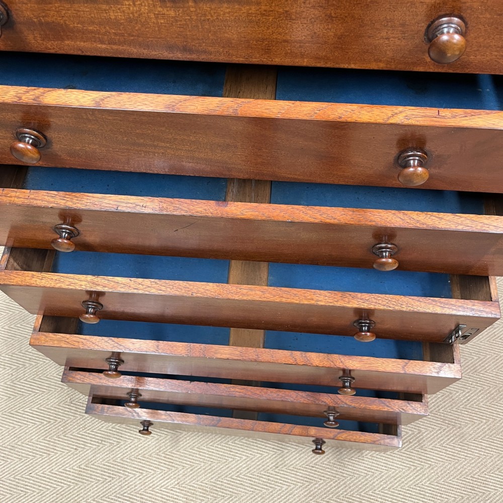 A tall Victorian mahogany Wellington chest featuring eight graduated drawers with turned wooden knobs, carved corbels at the top, and a side hinged locking flap on a plinth base. View IV