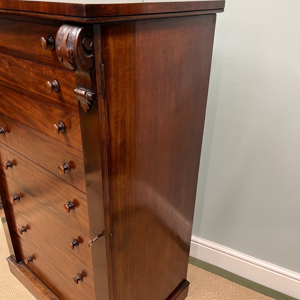 A tall Victorian mahogany Wellington chest featuring eight graduated drawers with turned wooden knobs, carved corbels at the top, and a side hinged locking flap on a plinth base. View X