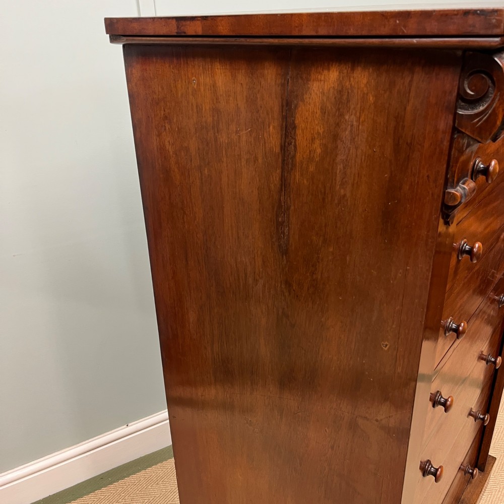 A tall Victorian mahogany Wellington chest featuring eight graduated drawers with turned wooden knobs, carved corbels at the top, and a side hinged locking flap on a plinth base. View XI