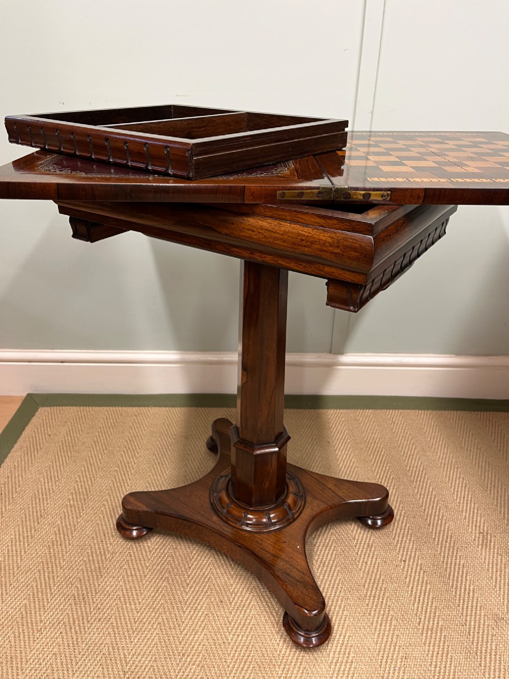 A 19th-century rosewood games table with a polished rectangular fold-over top, a faceted pedestal column, and a quadform base on flattened bun feet. View X