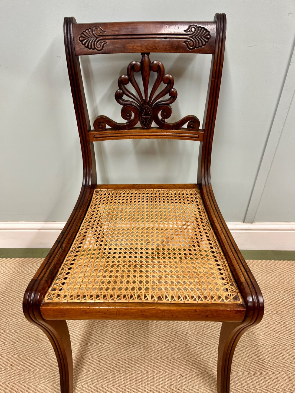 A set of six Regency carved rosewood dining chairs featuring carved anthemion back splats, sabre legs, and woven cane seats. View V