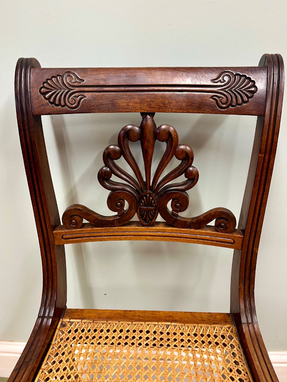 A set of six Regency carved rosewood dining chairs featuring carved anthemion back splats, sabre legs, and woven cane seats. View VI