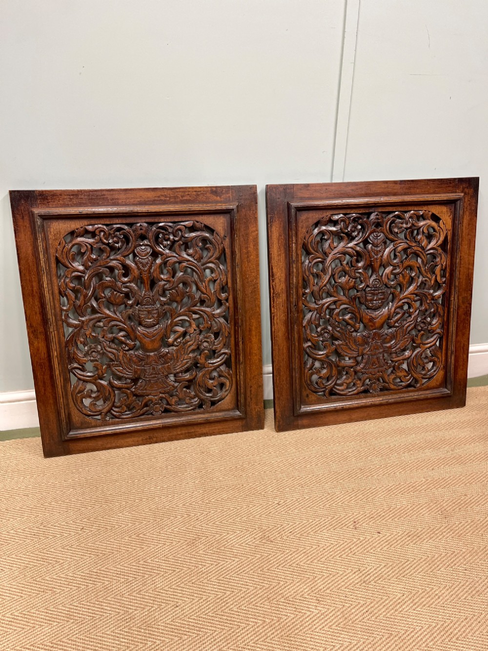 A pair of square dark teak wall panels featuring openwork floral and deity carvings surrounded by dark timber frames.