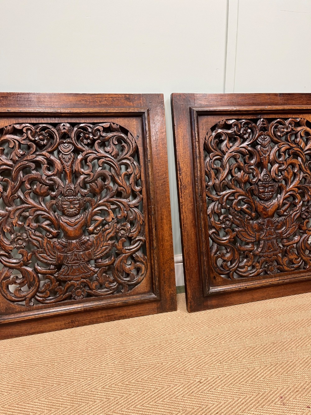 A pair of square dark teak wall panels featuring openwork floral and deity carvings surrounded by dark timber frames. View II