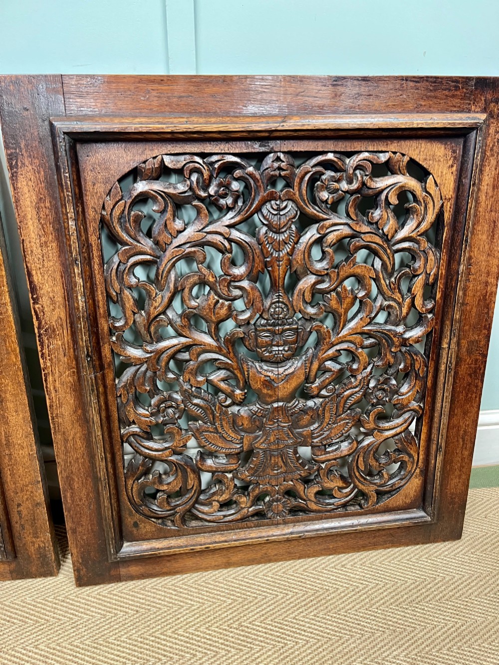 A pair of square dark teak wall panels featuring openwork floral and deity carvings surrounded by dark timber frames. View III